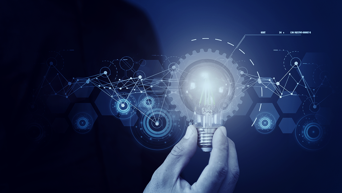 A person’s hand holds a lightbulb surrounded by a gear wheels and technology graphics representing ideas for technological innovation